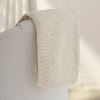 Cream knit throw blanket draped over the back of a neutral rounded-back chair.